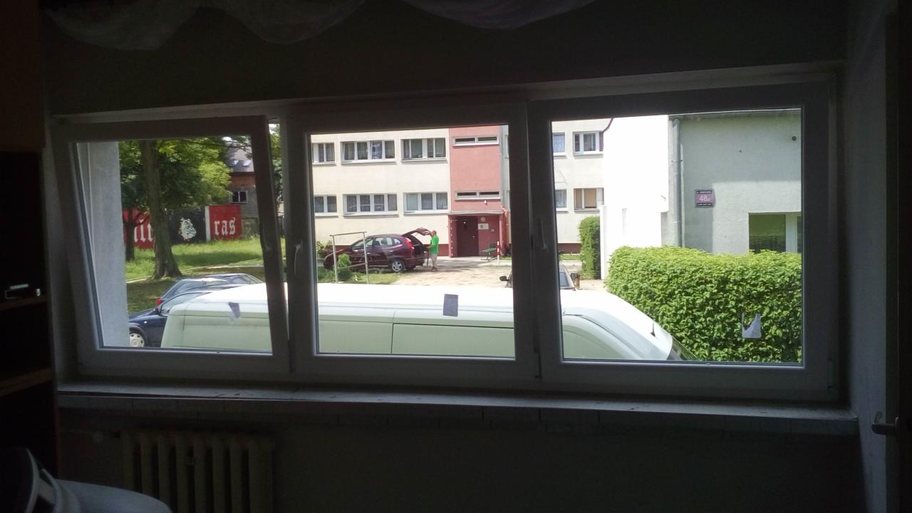 Trzyszybowe okno z widokiem na podwórko z zaparkowanymi samochodami i blokiem mieszkalnym w tle, widoczne firanki w górnej części ramy okiennej.