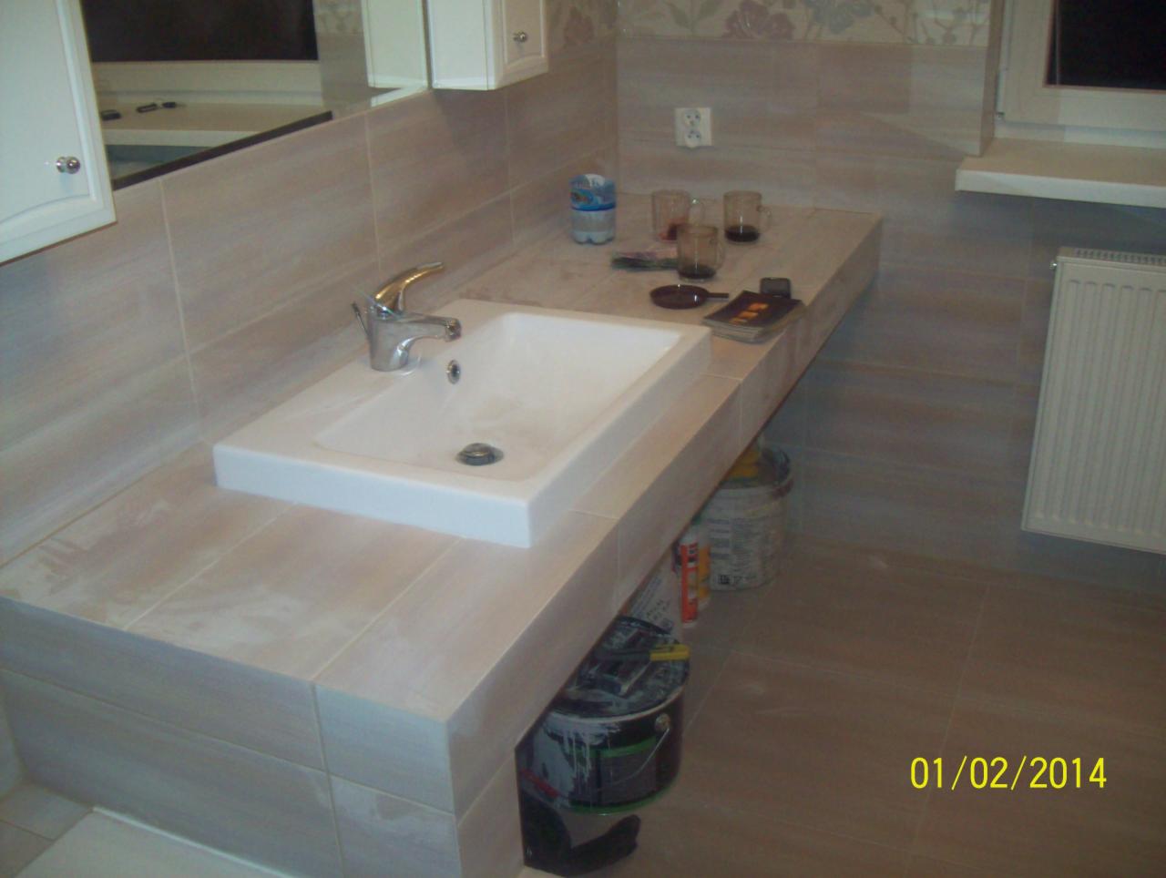 Wykończona łazienka z umywalką wpuszczoną w blat z płytek. Widoczne akcesoria, grzejnik i fragment ściany z tapetą. Ujęcie z perspektywy.