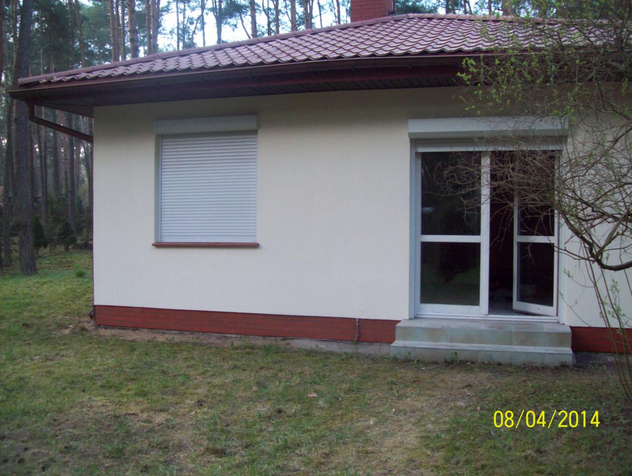 Niewielki, otynkowany dom z brązowym dachem i roletami zewnętrznymi, otoczony zielenią. Widoczny fragment elewacji z oknem i otwartymi drzwiami.