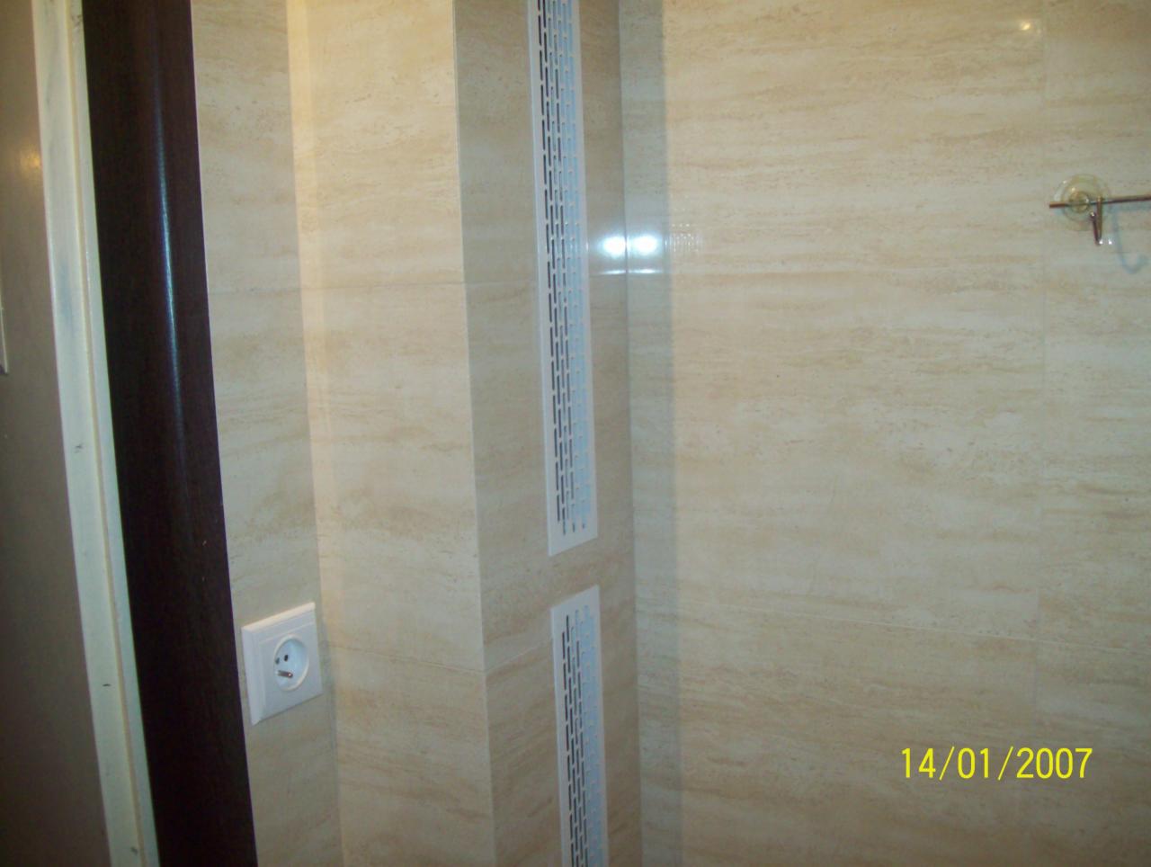 Beżowe płytki imitujące marmur w łazience w Ozorkowie. Widoczne kratki wentylacyjne i gniazdko elektryczne. Ciemne drzwi po lewej stronie kadru.