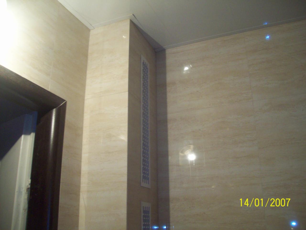 Wykończona łazienka z beżowymi płytkami imitującymi trawertyn, z widoczną kratką wentylacyjną w rogu i fragmentem ościeżnicy. Data w prawym dolnym rogu.