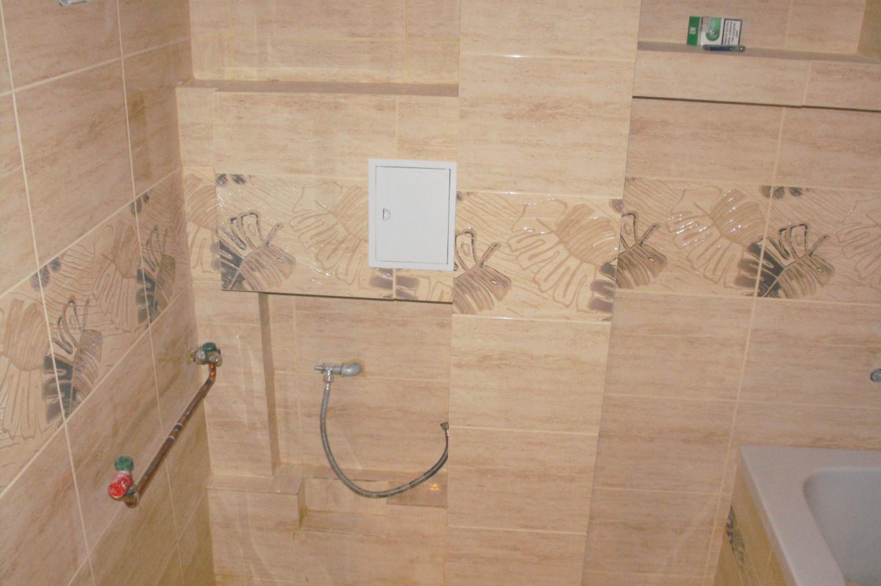 Beżowe płytki ścienne z motywem liści w łazience, widoczny zawór i biała klapka rewizyjna. Ujęcie z bliska.