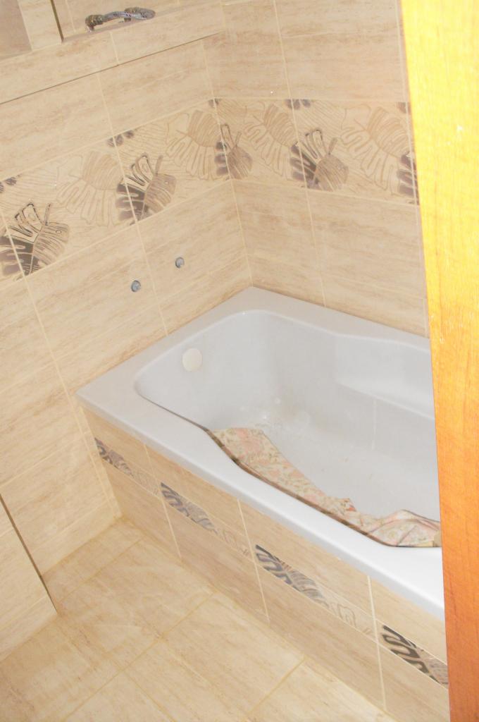 Jasna łazienka z beżowymi płytkami z motywem liści, biała wanna z zasłoną. Widok na fragment kabiny prysznicowej i drewnianej framugi drzwi.