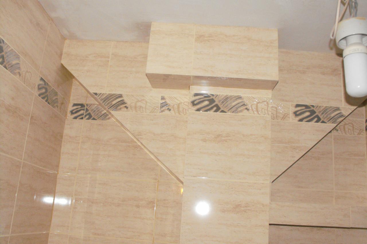 Wykończona łazienka z beżowymi płytkami imitującymi marmur, dekoracyjnym pasem z motywem liści i zabudową nad stelażem WC. Widoczne oświetlenie.