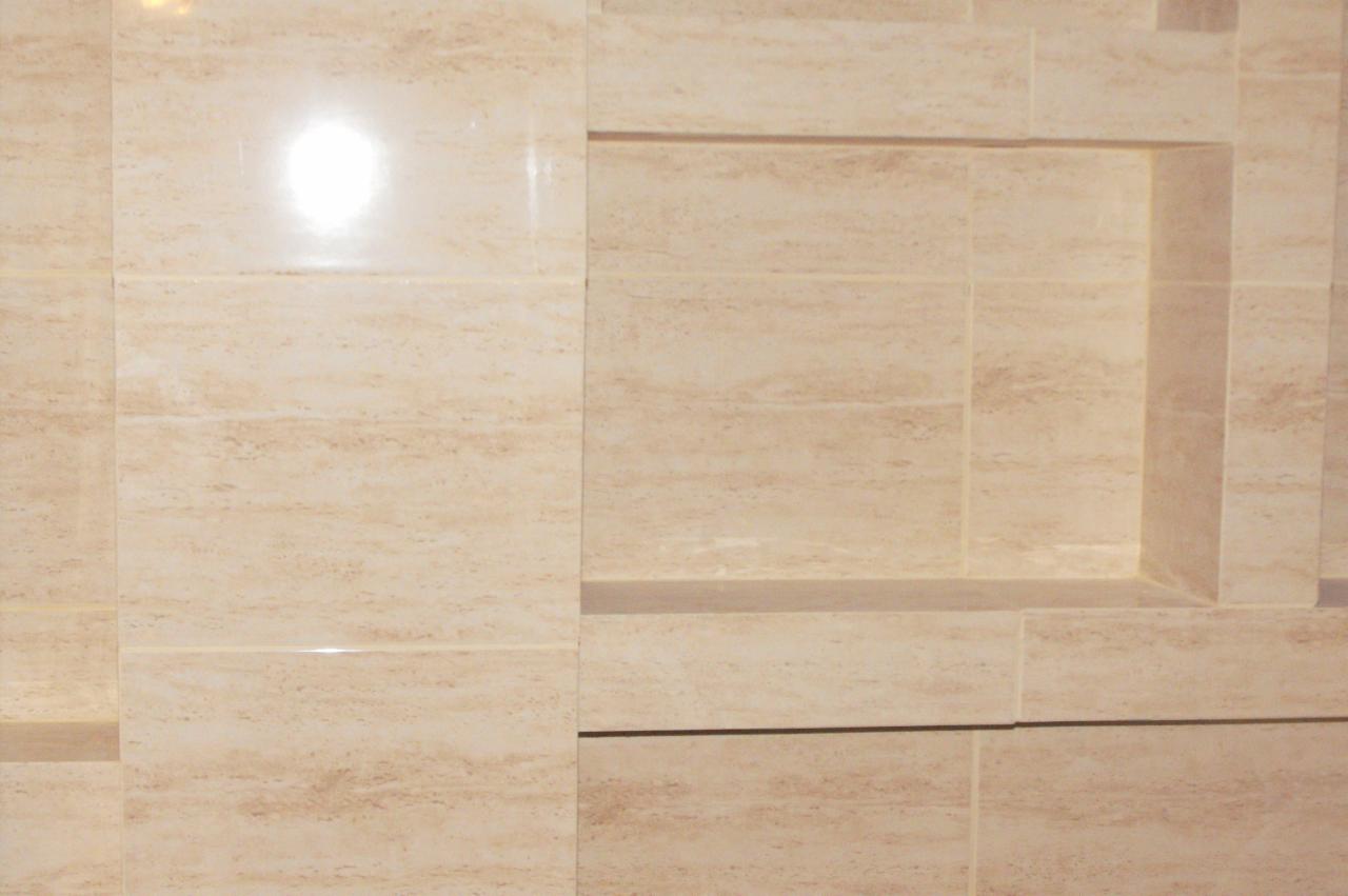 Beżowe, połyskujące płytki imitujące trawertyn, ułożone w łazience, z wnęką w ścianie. Widoczne fugi i odbicie światła na powierzchni płytek.