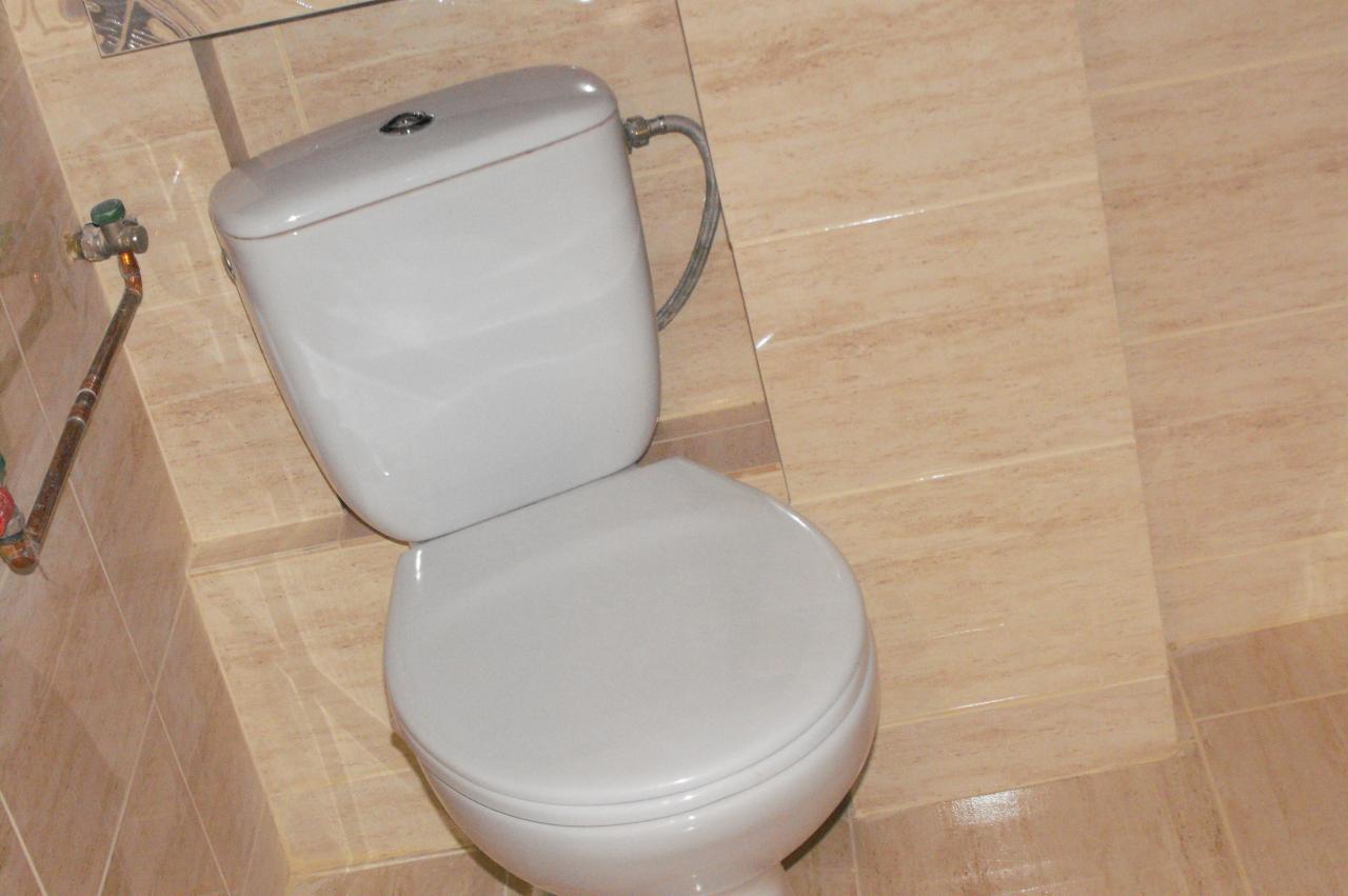 Biała toaleta na tle beżowych płytek ściennych i podłogowych w łazience po remoncie. Widoczne rury doprowadzające wodę. Ujęcie pod kątem.