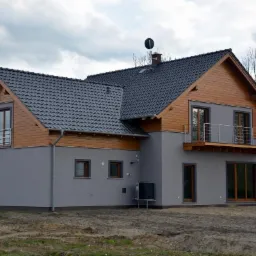 Dwukondygnacyjny dom z szarą elewacją i drewnianą górną częścią, balkon z metalową balustradą, ciemna dachówka, widoczny klimatyzator na zewnątrz.
