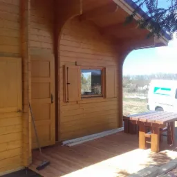 Wykończony drewniany domek z bali z zadaszonym tarasem, drewnianym stołem i ławkami, widoczne okiennice i detale architektoniczne, w tle biały samochód dostawczy.