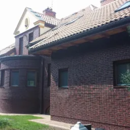 Dom z ciemnej cegły klinkierowej z jasną dachówką ceramiczną, widoczne rynny i fragment ogrodu z zielenią.