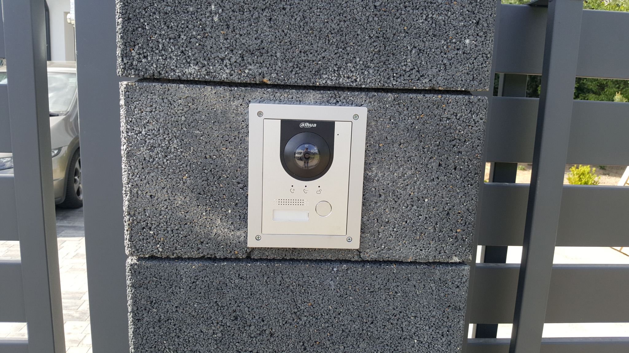 Stacja bramowa wideodomofonu DAHUA zamontowana na szarym, betonowym słupku ogrodzenia, widok z bliska przez szczeble metalowej bramy.
