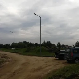 Dwie nowoczesne latarnie uliczne na skraju drogi gruntowej w Koziegłowach, z samochodami zaparkowanymi w pobliżu i pochmurnym niebem w tle.