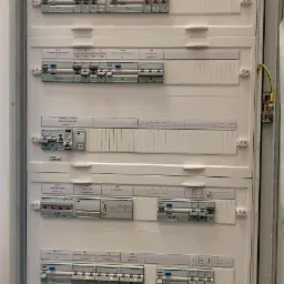 Rozdzielnica elektryczna z licznymi wyłącznikami nadprądowymi i różnicowoprądowymi, zabezpieczenia przepięciowe, opisana etykietami i schematami, widoczny przewód uziemiający.