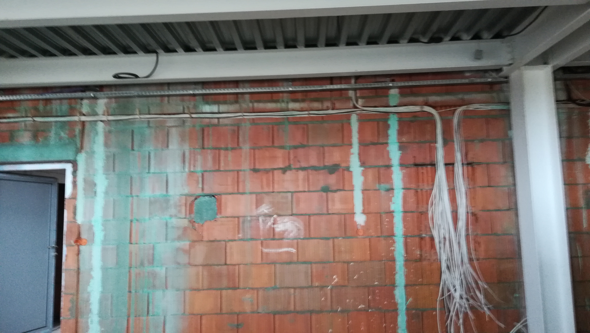 Ściana z cegły z widocznymi przewodami elektrycznymi w peszlach, korytka kablowe pod sufitem, ślady po wierceniu i zielone plamy po hydroizolacji.