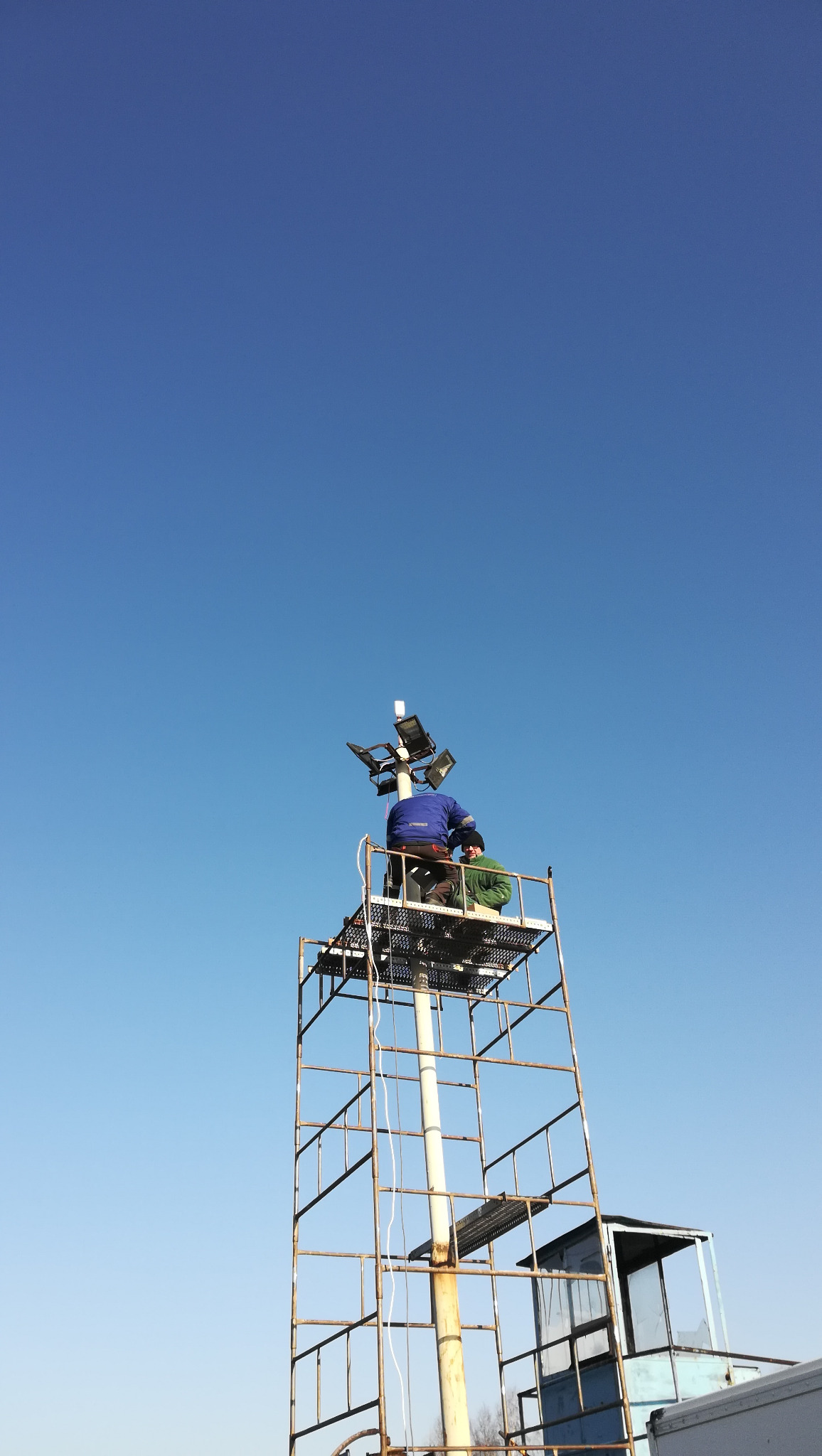 Dwóch monterów na metalowej konstrukcji wieży oświetleniowej, instalujących reflektory pod błękitnym niebem.