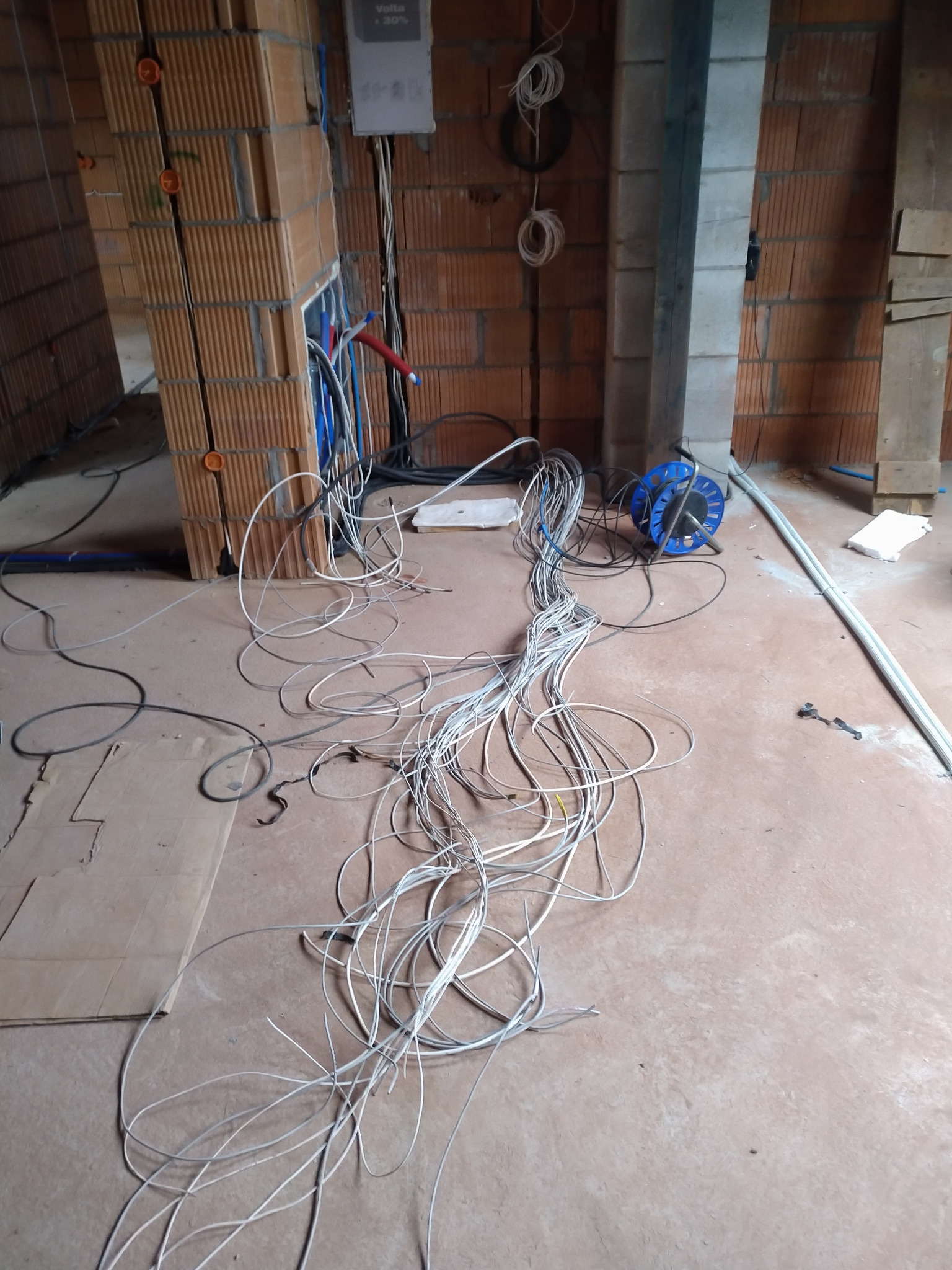 Surowy stan instalacji elektrycznej w budynku w trakcie budowy: kable zwisają z muru z cegły, a ich nadmiar leży na podłodze obok niebieskiego zwijadła.