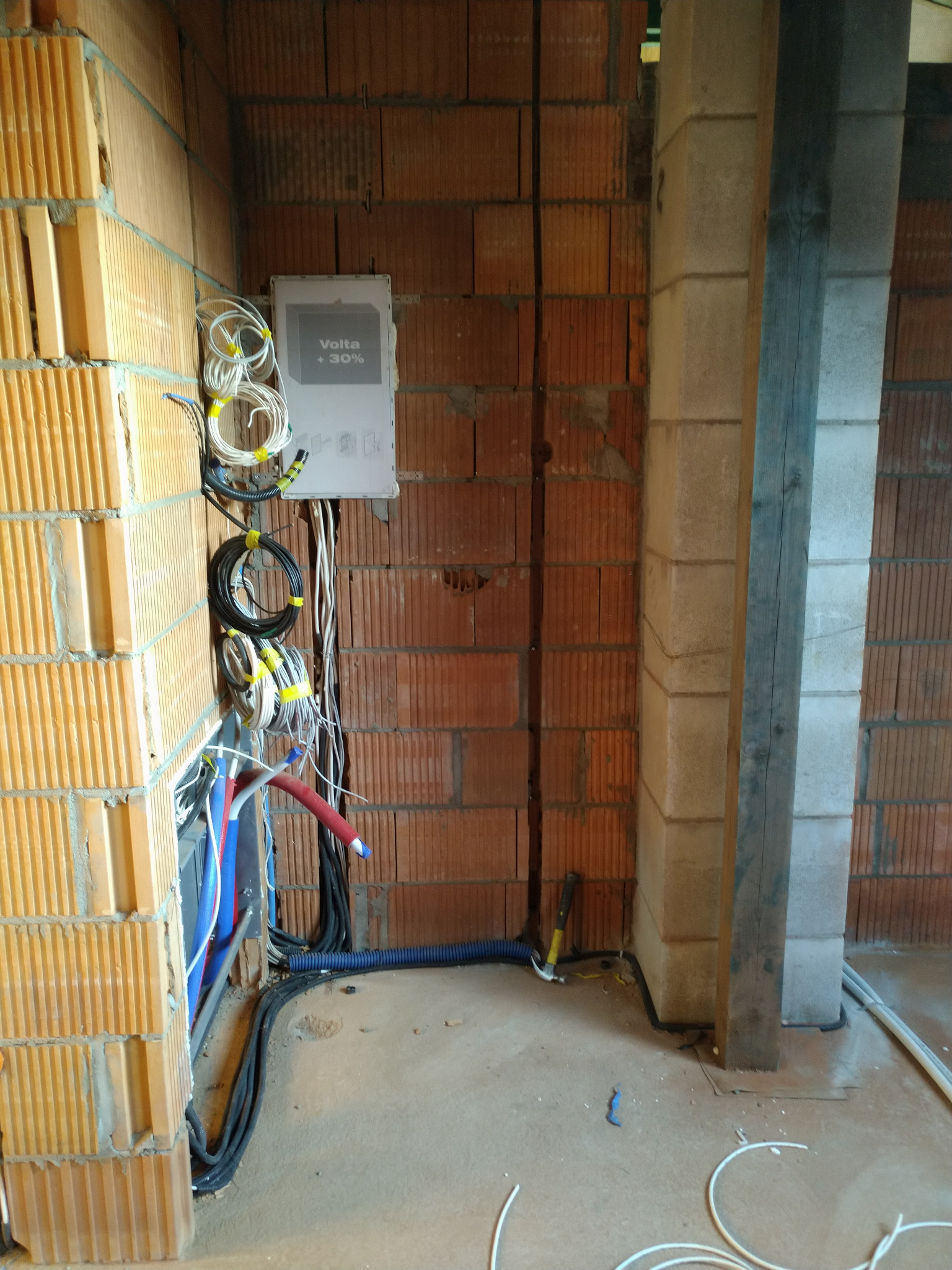 Wnętrze w trakcie prac instalacyjnych: rozdzielnia elektryczna Volta +30% na ceglanej ścianie, wiązki przewodów zabezpieczone żółtą taśmą, rury instalacyjne w kolorach niebieskim i czerwonym...