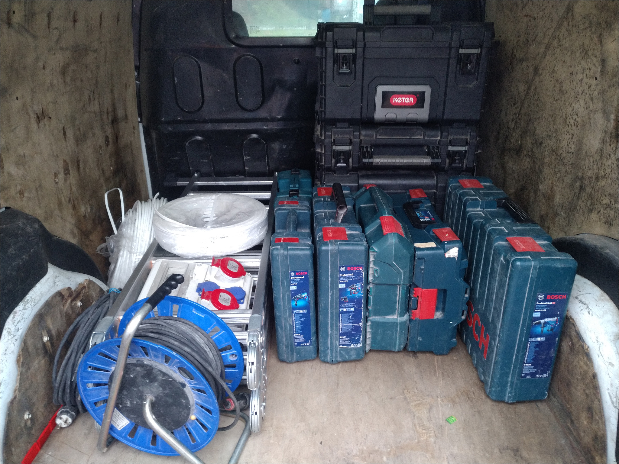 Wnętrze furgonetki wypełnione skrzynkami narzędziowymi marki Bosch, zwiniętym kablem na bębnie, drabiną aluminiową i rolką białego przewodu elektrycznego.