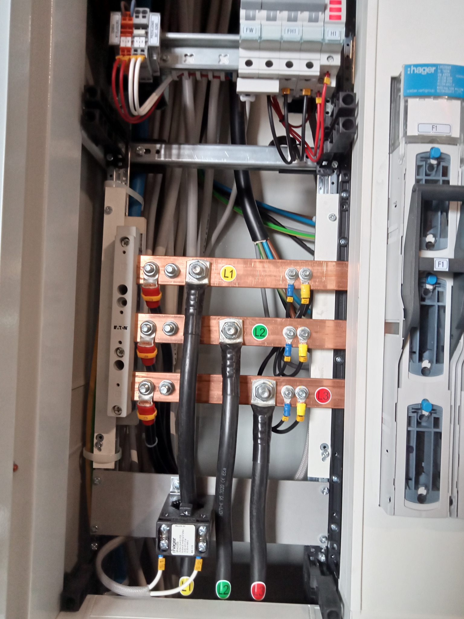 Wnętrze rozdzielnicy elektrycznej z widocznymi szynami zbiorczymi oznaczonymi L1, L2, L3, różnymi przewodami, bezpiecznikami i modułem Hager. Zastosowano elementy Eaton.