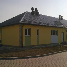 Żółta elewacja budynku z szarą dachówką i kominami. Fragment ocieplenia i malowania elewacji widoczny. Kostka brukowa na pierwszym planie.