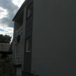 Elewacja budynku wielorodzinnego w odcieniach szarości, z balkonem i anteną satelitarną. Widok z dołu, częściowo zacieniony, z fragmentem dachu sąsiedniego budynku.