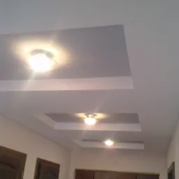 Wykończony sufit podwieszany z oświetleniem punktowym w salonie. Widoczne trzy kwadratowe wnęki z zamontowanymi lampami. Ściany w jasnym odcieniu.