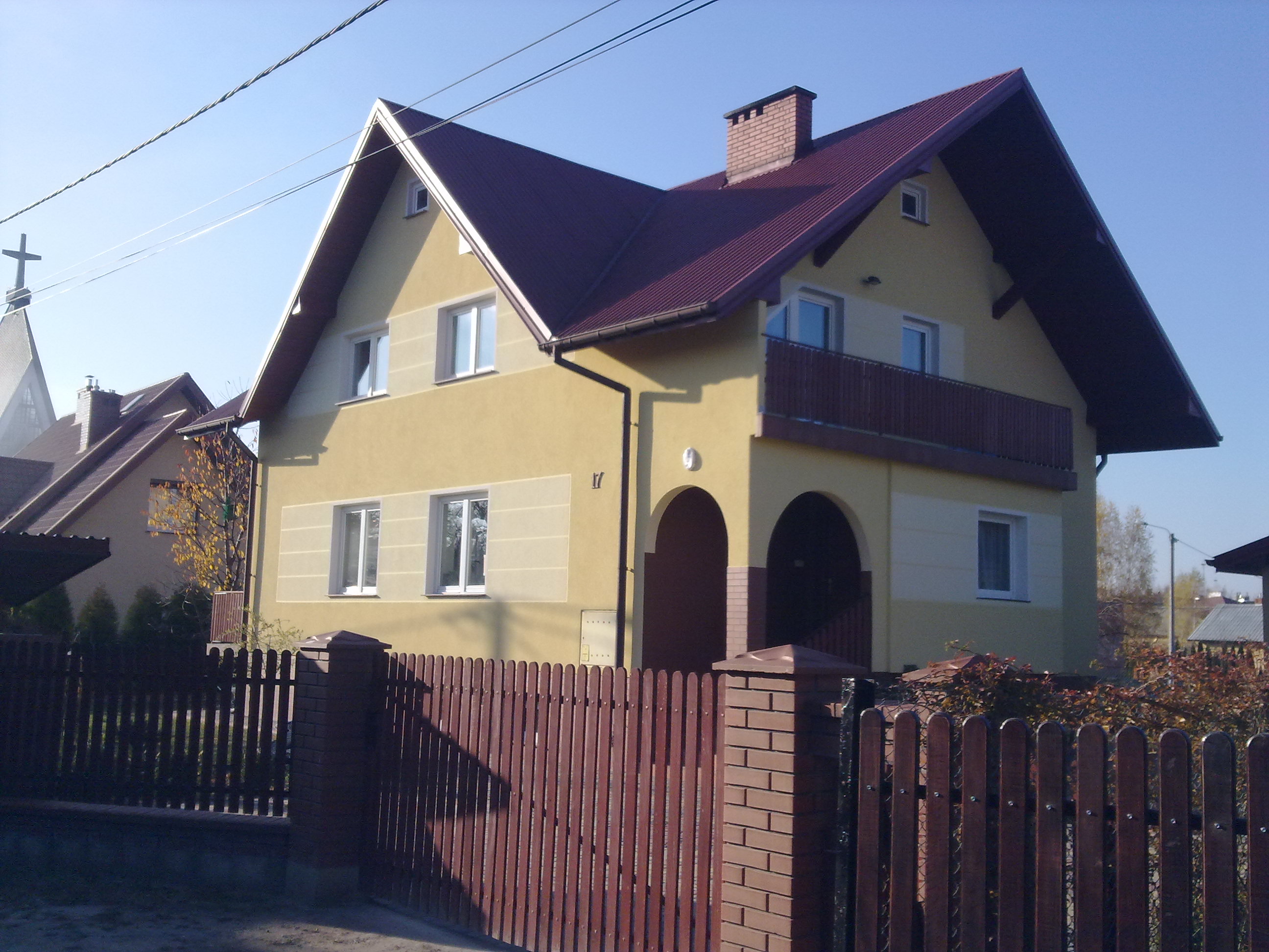 Dwukondygnacyjny dom z balkonem i czerwonym dachem, widoczny zza drewnianego ogrodzenia. Żółta elewacja, łukowate wejście. W tle fragment kościoła.