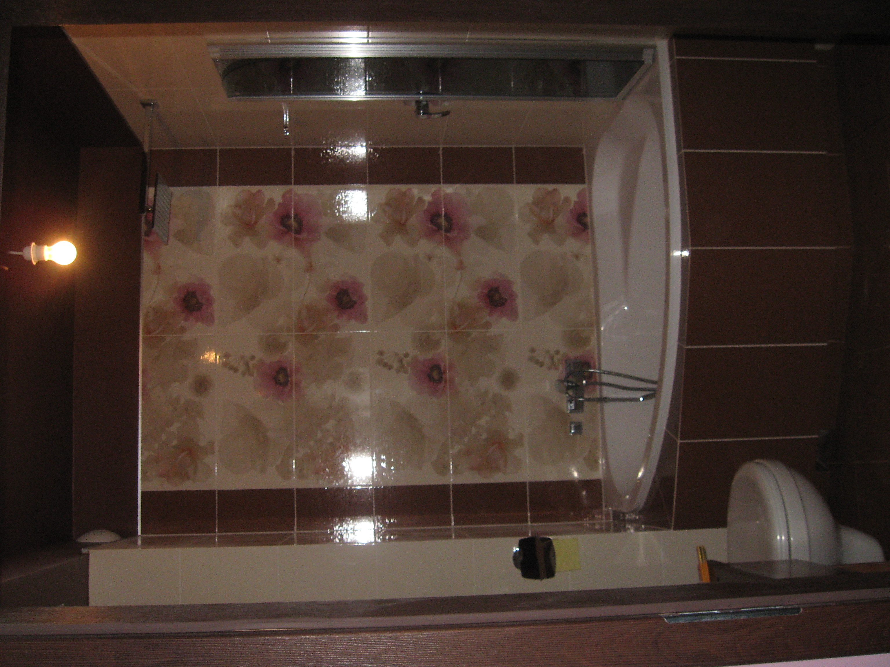 Wykończona łazienka z brązowo-kwiatowymi płytkami, białą wanną i toaletą. Widoczne oświetlenie i elementy armatury. Całość w ciepłej tonacji.