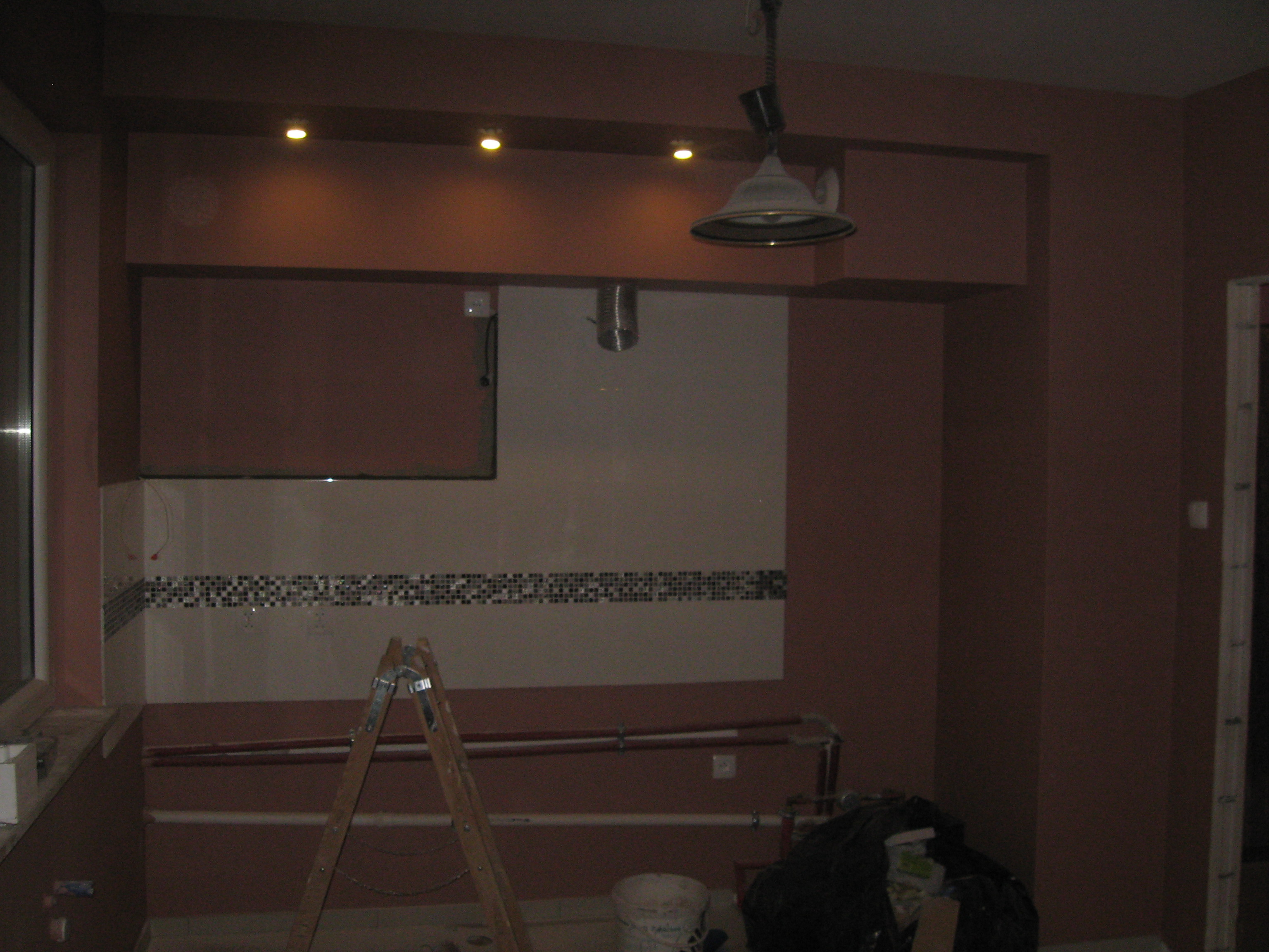 Remont kuchni w trakcie: ściany pomalowane na brązowo, częściowo ułożone płytki, drabina, rury i lampa wisząca. Widoczny etap prac wykończeniowych.