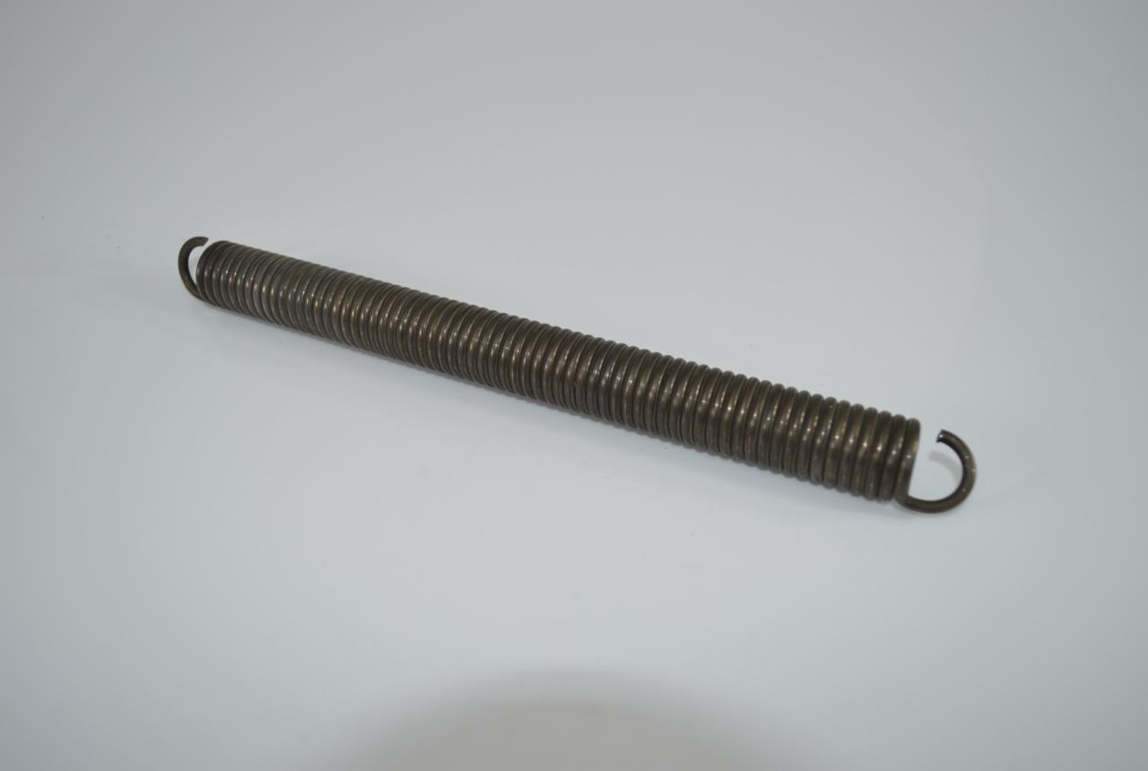 Metalowa sprężyna naciągowa z zakrzywionymi końcami, leżąca ukośnie na gładkim, jasnym tle. Widoczne detale zwojów i ich połączeń.