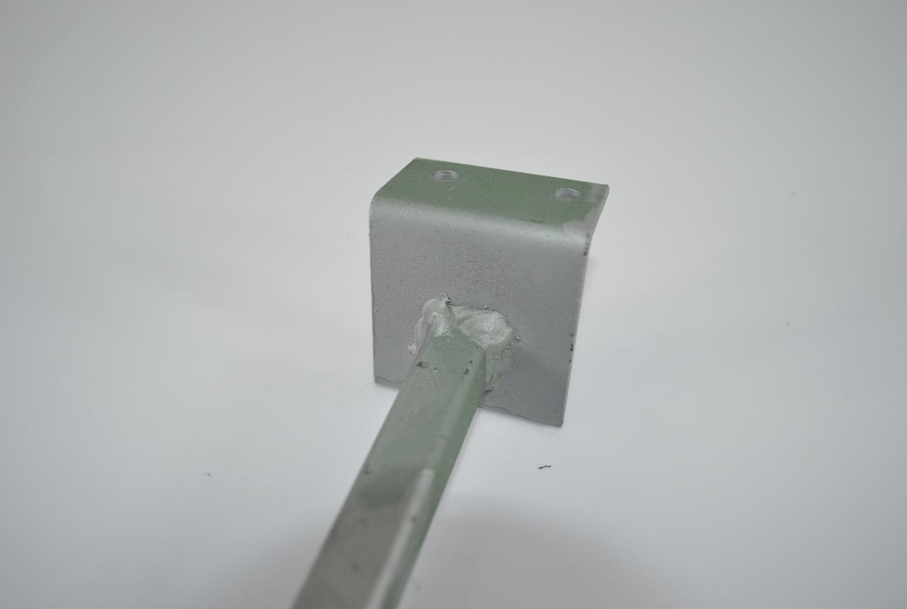 Szara, spawana konstrukcja metalowa z kątownika i pręta na białym tle. Widoczne ślady spawania i dwa otwory w kątowniku.