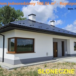 HETMAN Usługi Budowlane Jan Gazda Domy na sprzedaż - Nowoczesny parterowy dom jednorodzinny z ciemną dachówką, białymi ścianami i szarym cokołem, z brązowymi ramami okiennymi, widziany z zewnątrz w słoneczny dzień.