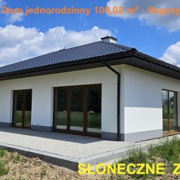 HETMAN Usługi Budowlane Jan Gazda Domy na sprzedaż - Nowoczesny, parterowy dom jednorodzinny z białymi ścianami, grafitowym dachem i brązowymi oknami, otoczony kostką brukową i zielenią.