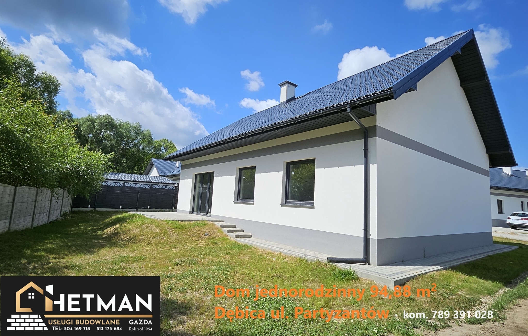 Nowoczesny dom jednorodzinny z ciemnoszarą dachówką i białymi ścianami, widoczny z zewnątrz w słoneczny dzień, z logo firmy budowlanej Hetman w lewym dolnym rogu.