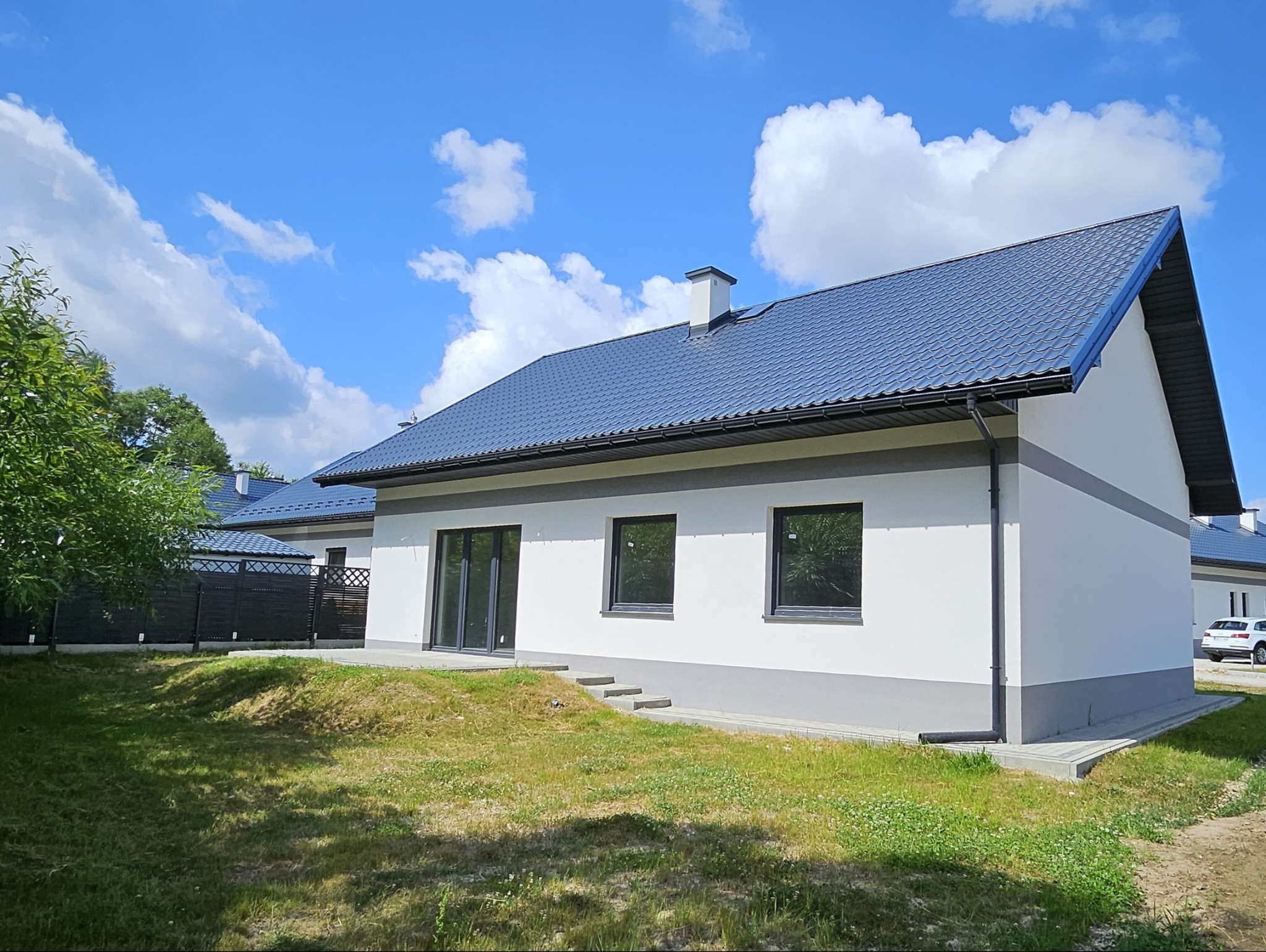Świeżo wybudowany, parterowy dom z ciemną dachówką i białymi ścianami, widoczny z boku, z fragmentem ogrodu i błękitnym niebem w tle.