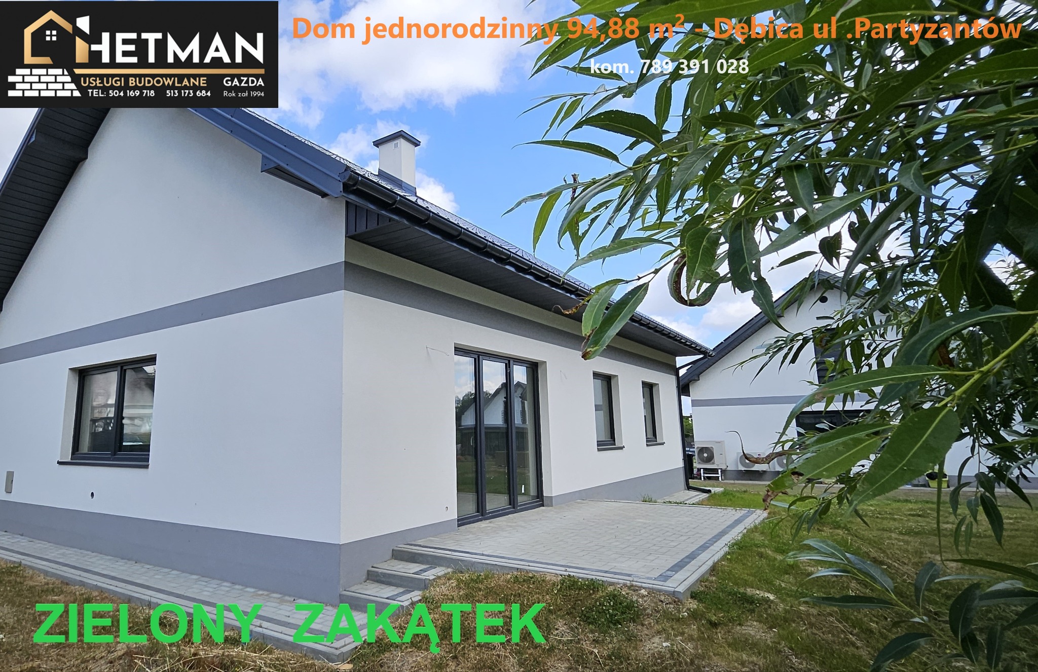 Nowoczesny dom jednorodzinny z szarą podmurówką i obramowaniami okien, widok z boku na taras wyłożony kostką, zieleń w tle i logo firmy budowlanej w górnym rogu.