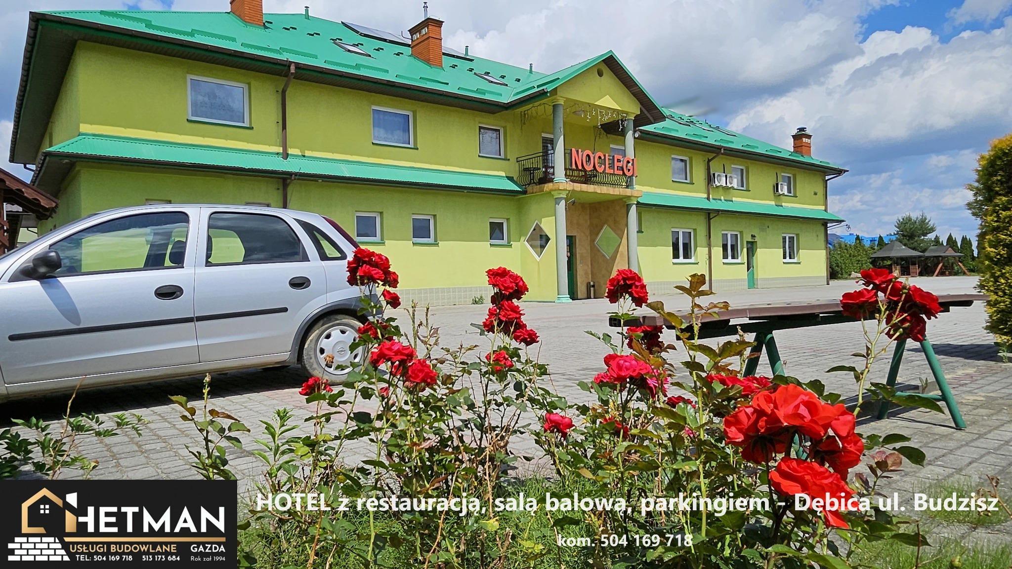 Żółto-zielony budynek z napisem 'NOCLEGI' widziany z perspektywy ogrodu z kwitnącymi czerwonymi różami i zaparkowanym srebrnym samochodem.