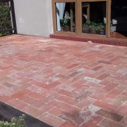 Ceglany taras ułożony w jodełkę, przylegający do domu z oknem tarasowym i fragmentem balustrady.