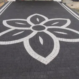 Czarno-biała nawierzchnia z kostki z motywem kwiatowym, wzór ułożony z drobnych kostek w kształcie wachlarza, białe linie obramowują obszar, w tle widoczny sprzęt budowlany.