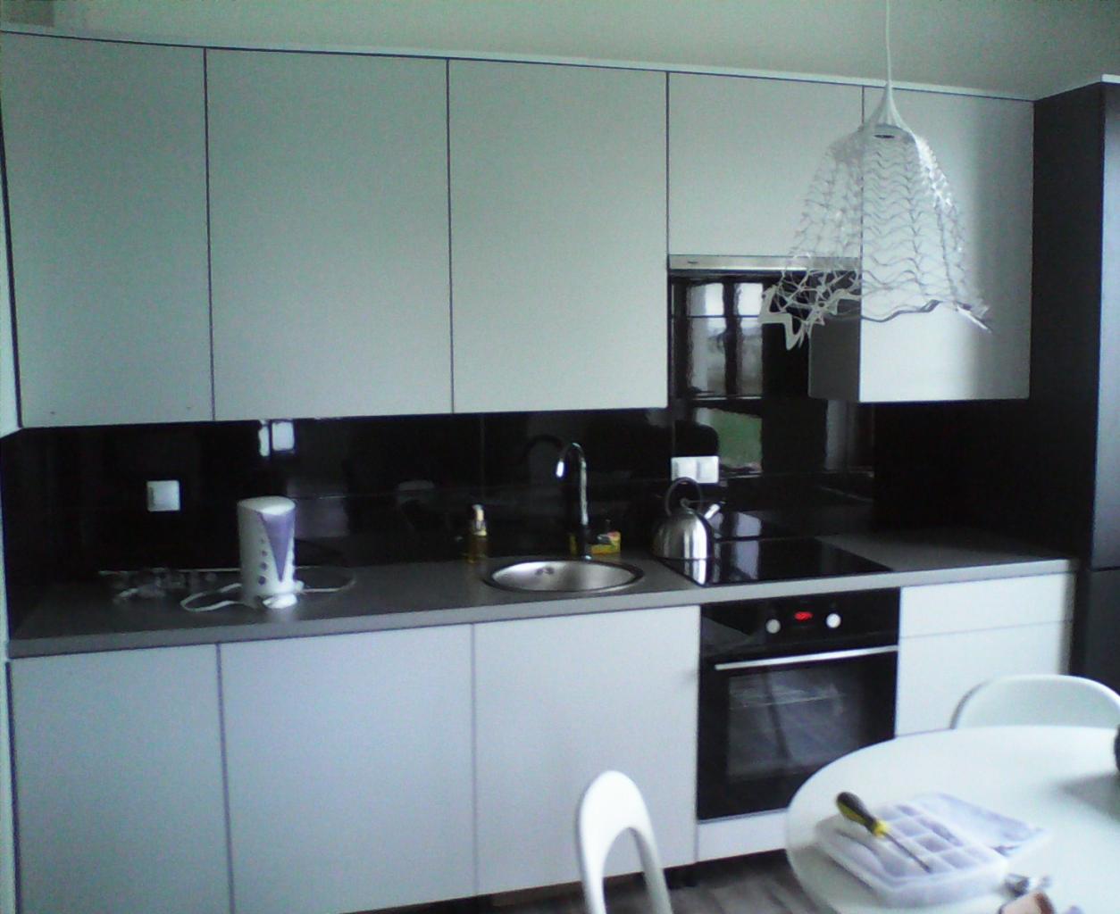 Nowoczesna kuchnia z białymi, matowymi frontami, czarnym panelem nad blatem i designerską lampą. Na blacie czajnik i drobne akcesoria kuchenne.