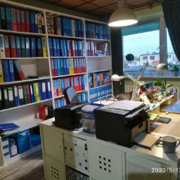 Biuro z regałem wypełnionym segregatorami, biurkiem z laptopem, lampkami i drukarkami, widok z perspektywy osoby stojącej w pomieszczeniu.