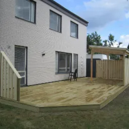 Jasny, drewniany taras przylegający do dwukondygnacyjnego domu z białej cegły, z drewnianą pergolą i częściowym ogrodzeniem. Widok z poziomu trawnika.