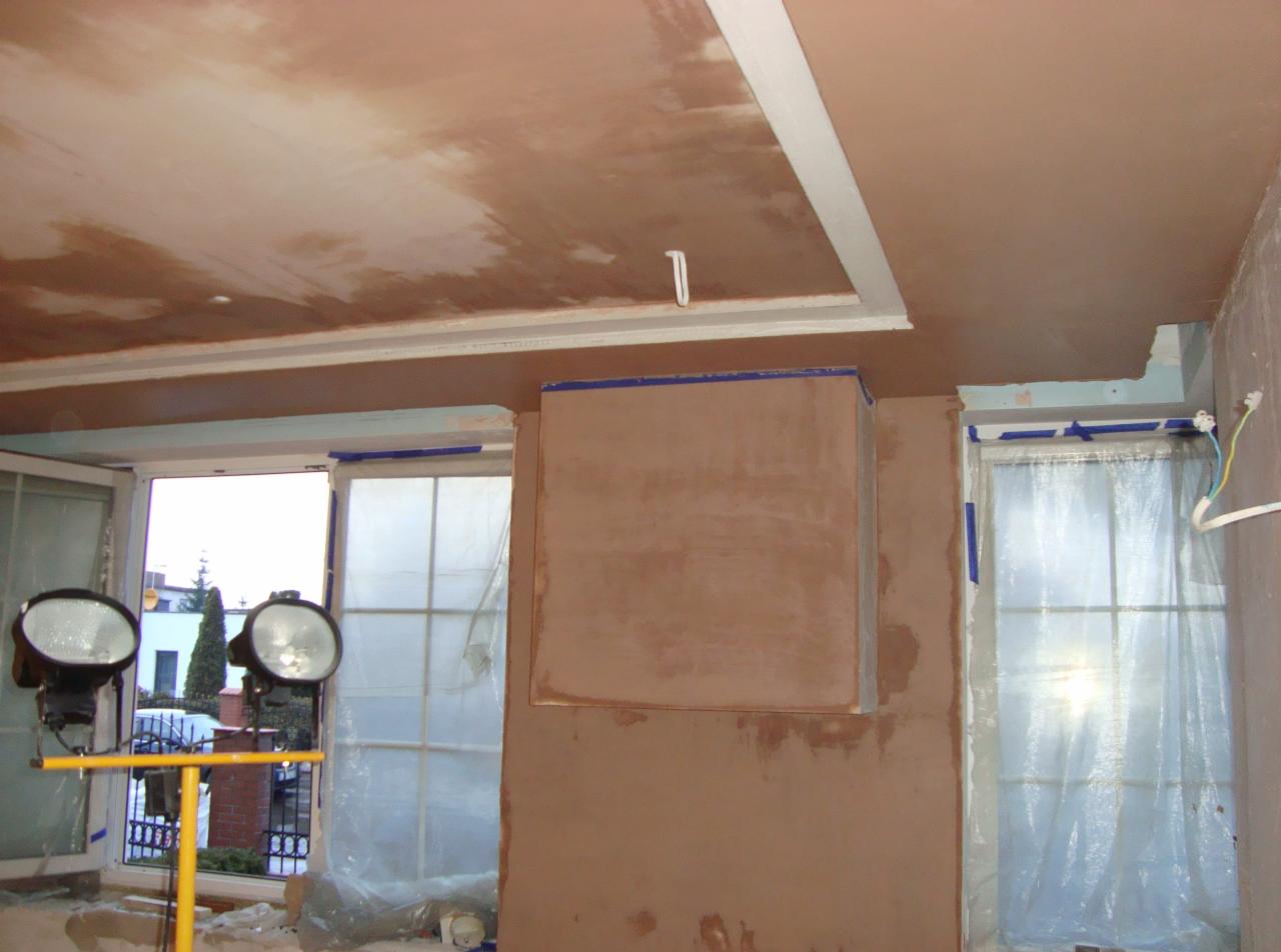Ściana i sufit w trakcie szpachlowania, zabezpieczone okna folią, widoczne lampy budowlane. Prace wykończeniowe w pomieszczeniu z oknami.