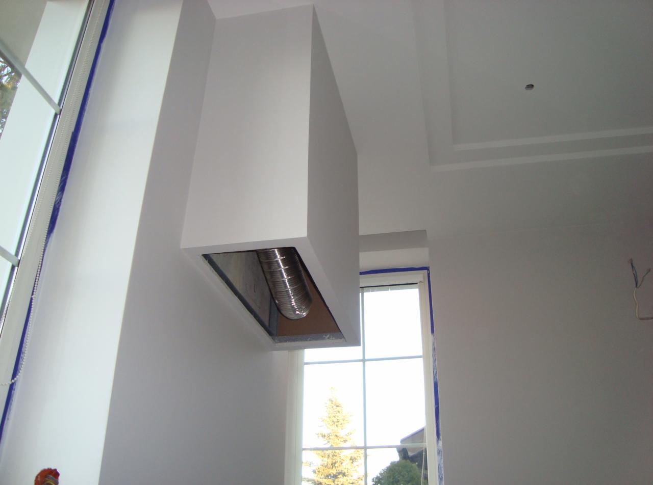Biały okap kuchenny z widocznym kanałem wentylacyjnym na tle okna i świeżo pomalowanej ściany. Widoczne zabezpieczenia okna folią malarską.