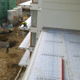 Montaż izolacji na balkonach budynku wielorodzinnego. Widoczne materiały izolacyjne Botament, pracownicy w odblaskowych kamizelkach i poziomica. Ujęcie z góry.