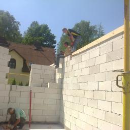 Usługi Remontowo-Budowlane Kołakowski Maciej - Wznoszenie ścian domu z białych bloczków, widoczni pracownicy. W tle budynek z brązowym dachem i drzewa. Żółta podpórka stabilizująca konstrukcję.