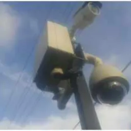 Zewnętrzna kamera obrotowa i skrzynka sterująca zamontowane na słupie na tle błękitnego nieba z chmurami i liniami energetycznymi. Ujęcie z dołu.