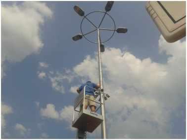 Pracownik w koszu podnośnika montuje lampy na wysokim maszcie oświetleniowym, na tle błękitnego nieba z chmurami. Widok z dołu.