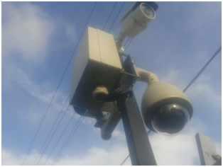 Zewnętrzna kamera obrotowa i skrzynka sterująca zamontowane na słupie na tle błękitnego nieba z chmurami i liniami energetycznymi. Ujęcie z dołu.