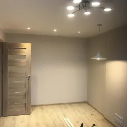 Wnętrze pokoju po remoncie z drewnianą podłogą, nową drewnianą stolarką drzwiową z przeszkleniami, nowoczesnym oświetleniem sufitowym i wiszącą lampą. Na podłodze leżą listwy przypodłogowe.