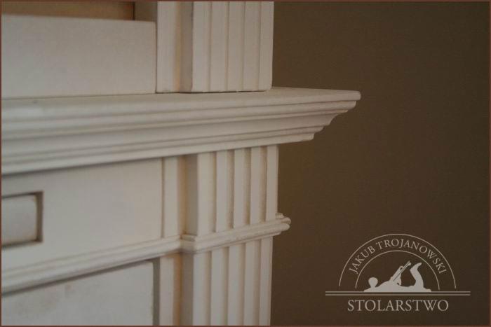 Fragment białej, drewnianej biblioteczki z dekoracyjnymi listwami i gzymsem, w tle logo firmy stolarskiej.